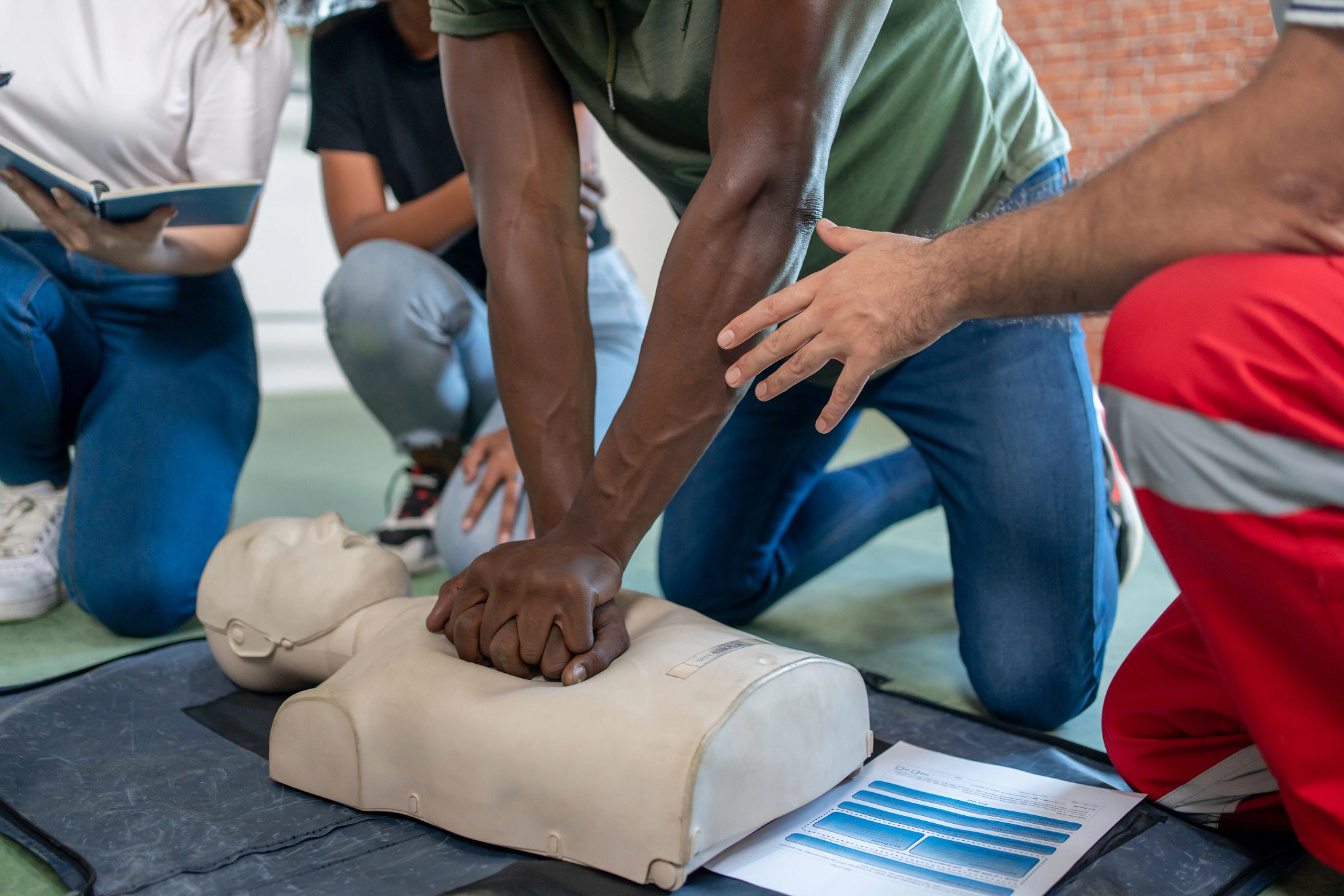 close-up of a person practicing chest compressions on a CPR mannequin during a training class with an instructor and students nearby