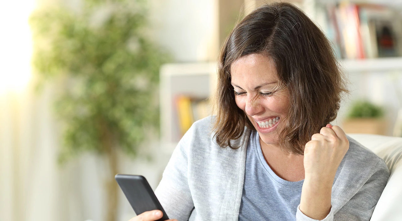 Excited adult woman celebrating good news on smart phone sitting on the couch at home