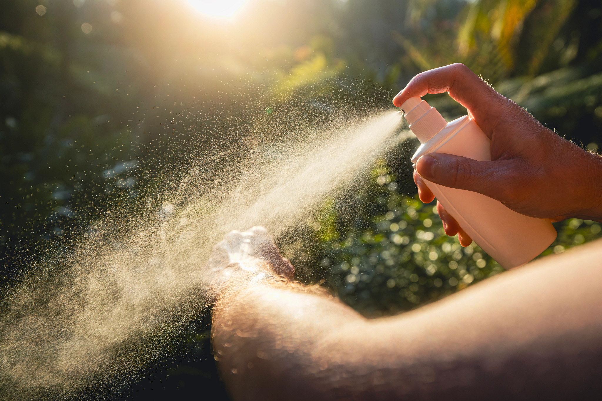 Close-up of a hand spraying a fine mist of insect repellent from a white bottle onto a person's forearm, with green foliage and bright sunlight in the background.