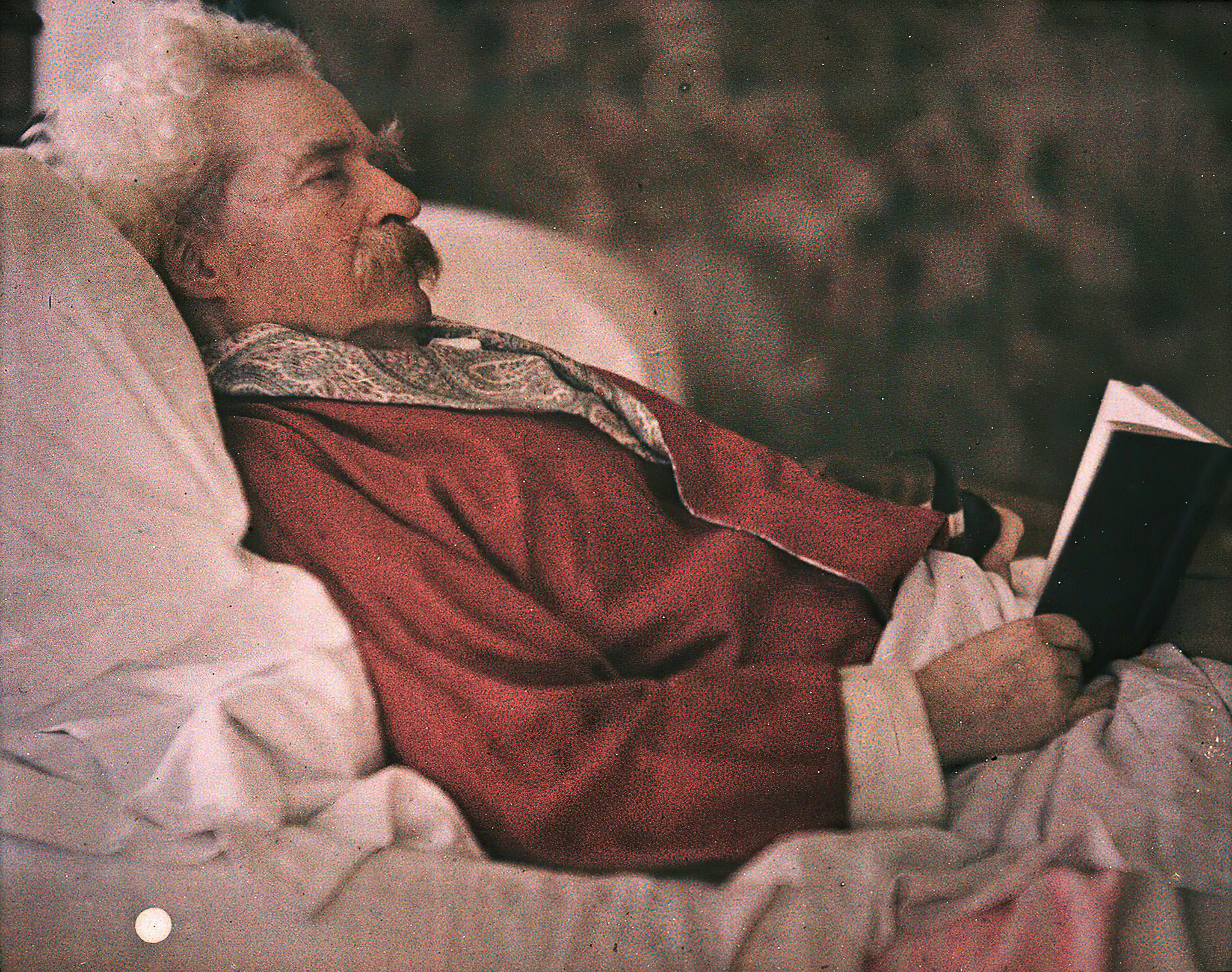 Mark Twain as an older man, reclining with a book and a tobacco pipe, wearing a red bed jacket