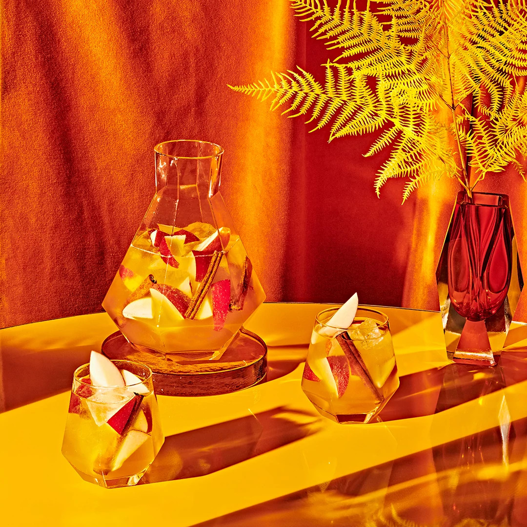 Glass carafe and two fruit-garnished mocktails arranged on a golden table with a fern backdrop