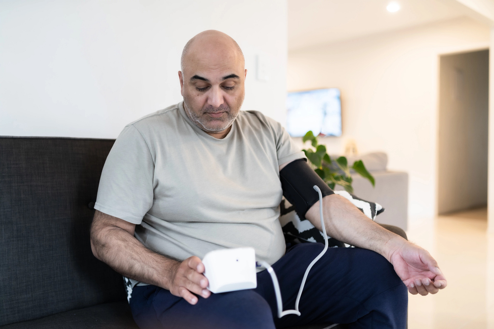 man with a shaved head sitting on a dark grey sofa, using a digital blood pressure monitor