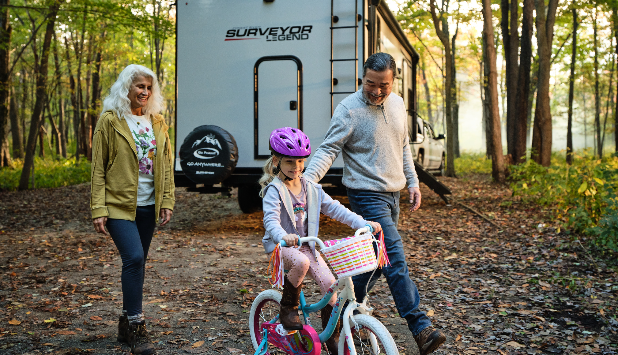 grandparents helping young daughter learn to ride a bike with a travel trailer in the background