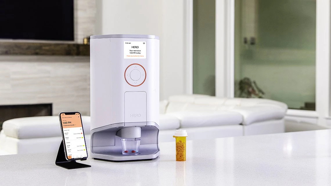 Hero Smart Pill Dispenser Hero Smart Pill Dispenser on counter top in living room with smart phone next to it and standard pill bottle with pills in it on counter top