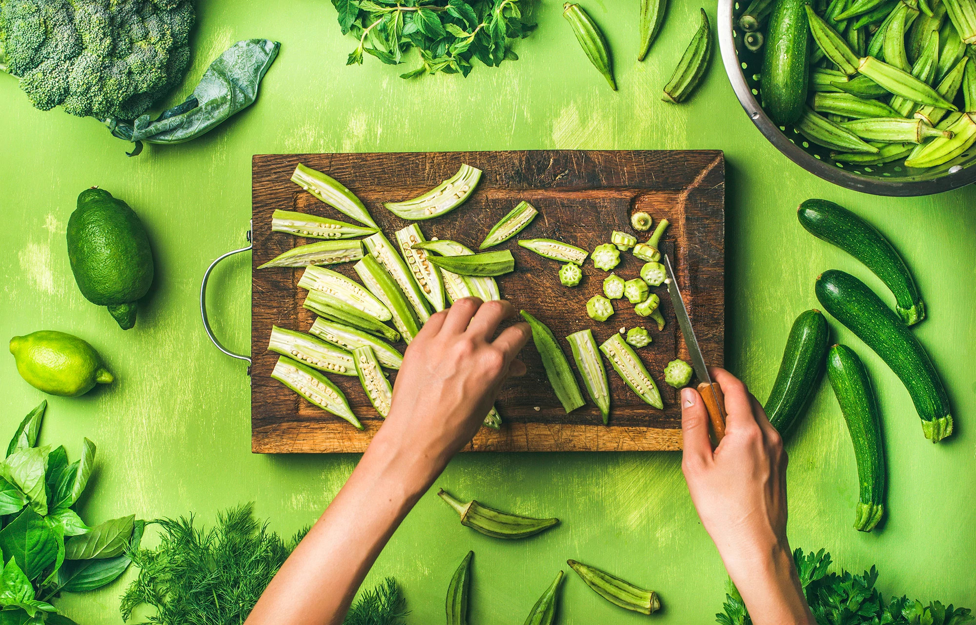 Woman's hands chopping zucchini on a cutting board surrounded by green veggies and background