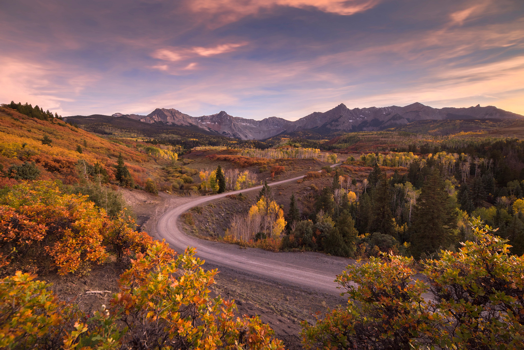 Scenic mountain landscape featuring a winding dirt road, colorful fall foliage, forests, and rugged mountain peaks under a soft sunset sky