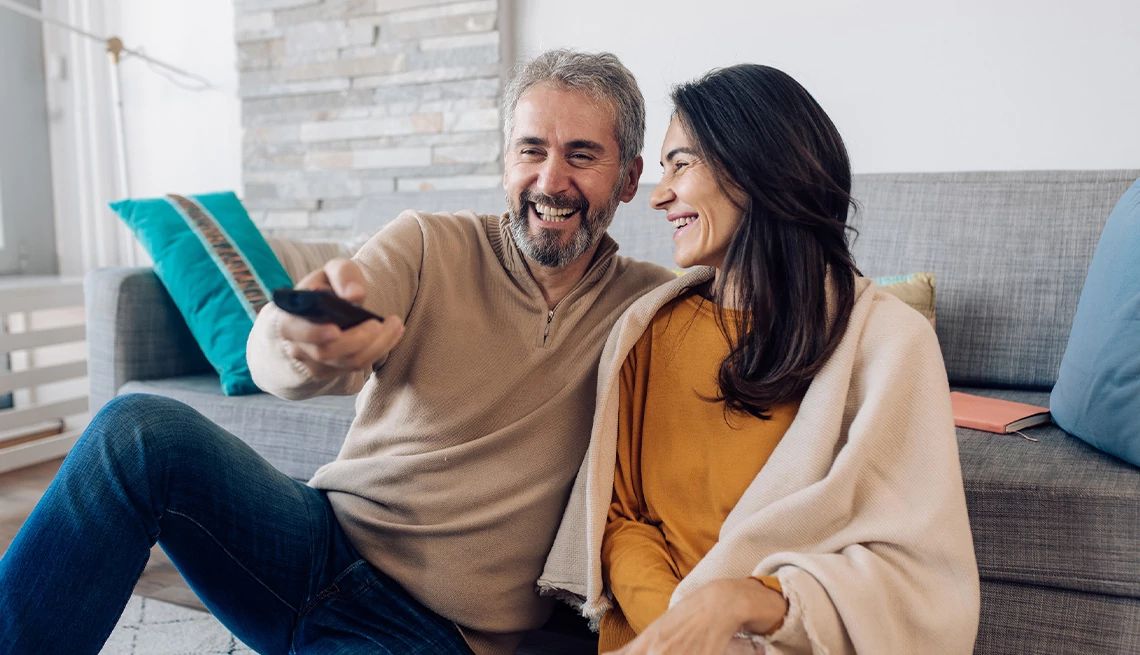 Couple relaxing in their living room in front of grey couch with man pointing TV remote to change the channel