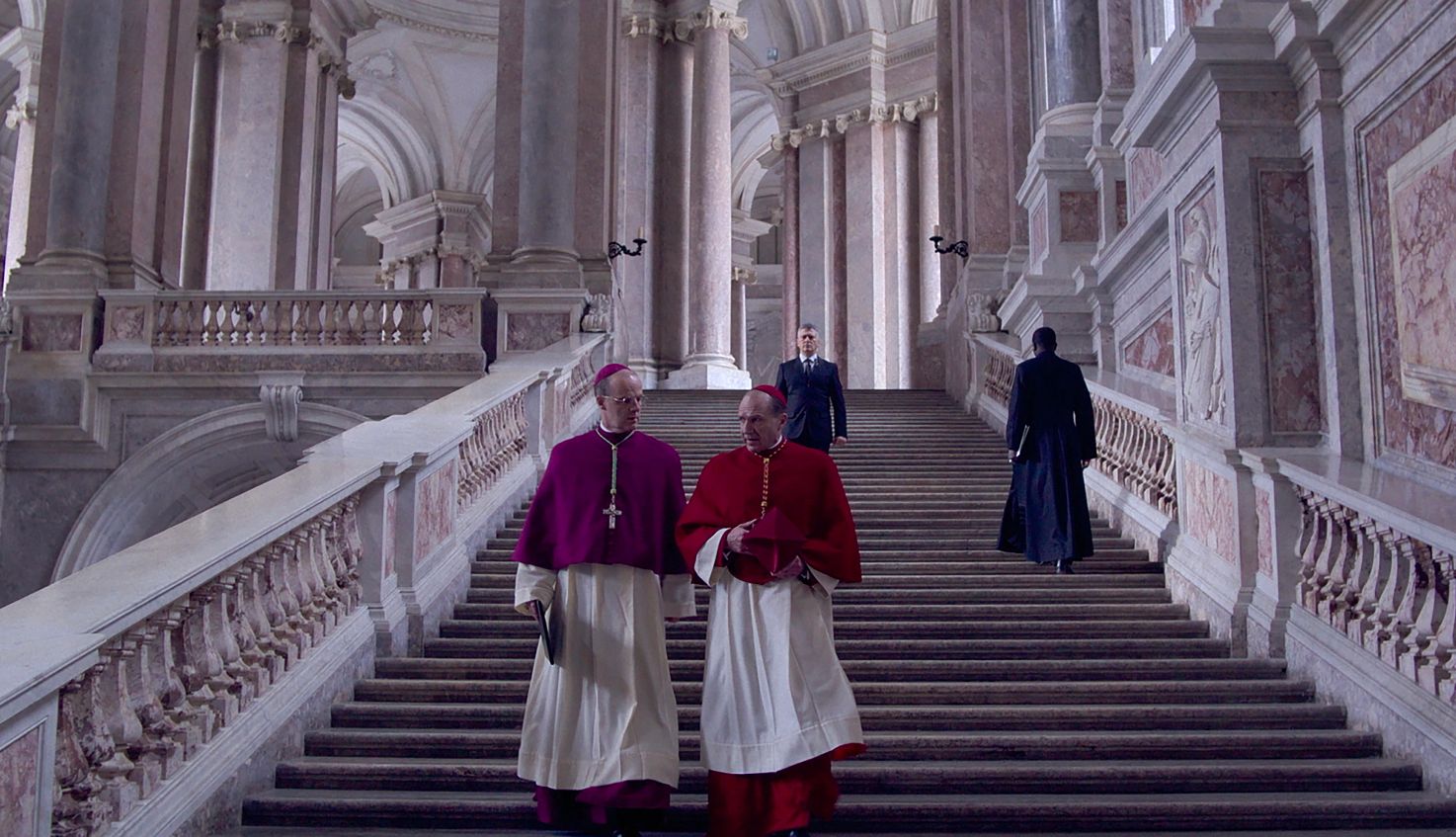 Conclave two men dressed as Cardinals walk down a large staircase in a scene from conclave movie