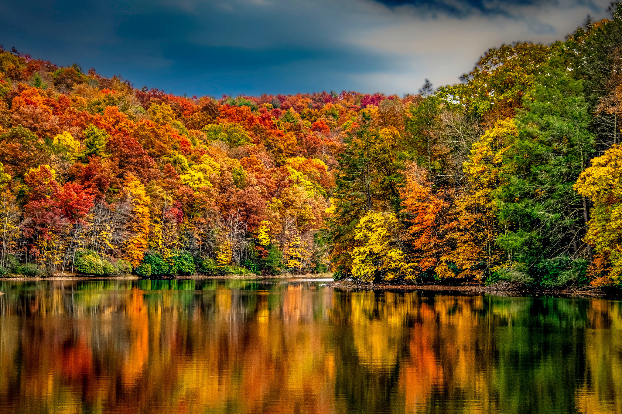 Scenic autumn landscape featuring a forest covered in colorful fall foliage reflected in a calm lake under a dramatic sky