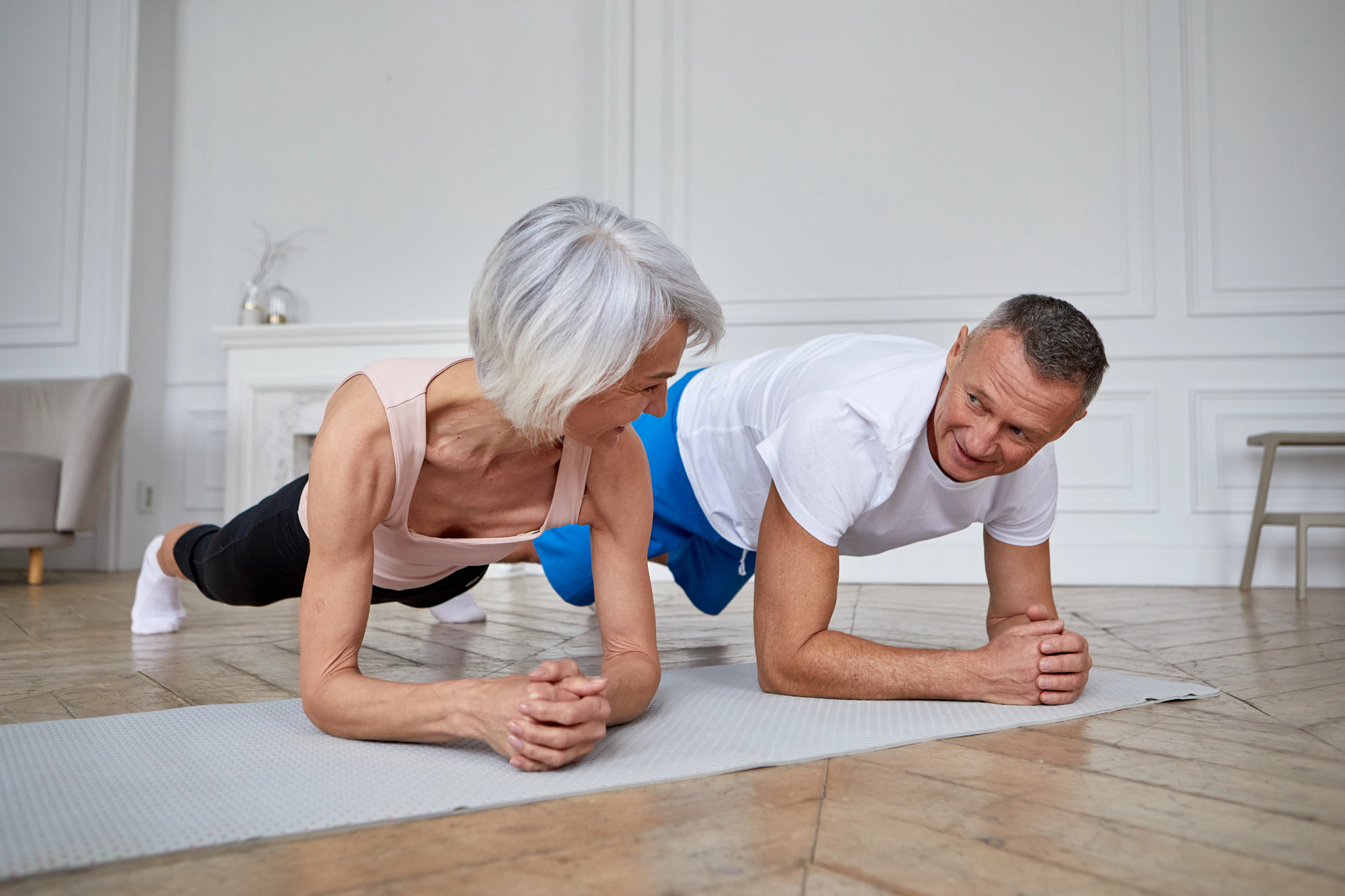 Positive athletic middle aged couple in sportswear doing plank exercise on mat and looking at each other while training together in light living room