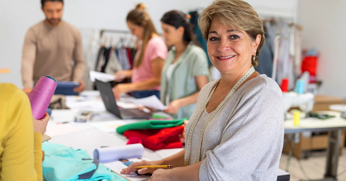 A person standing and smiling while sewing a garment in a sewing room with other people in the background sewing.