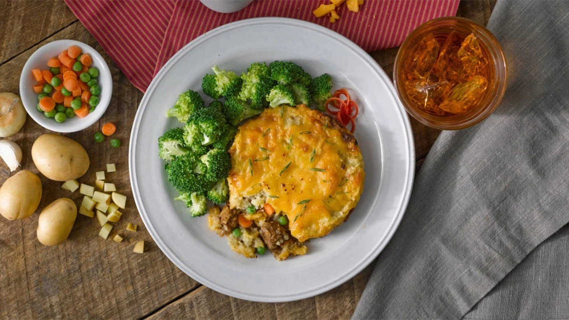 Shepherds pie with side of peas and carrots mix plus broccoli, plated on a wood table, ice tea drink