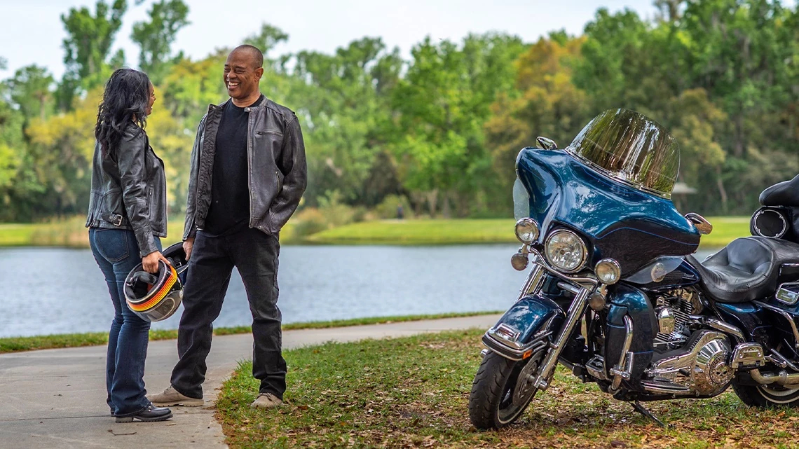 couple in leather jackets, pants, holding motorcycle helmets, standing by a lake on a concrete path, trees, grass