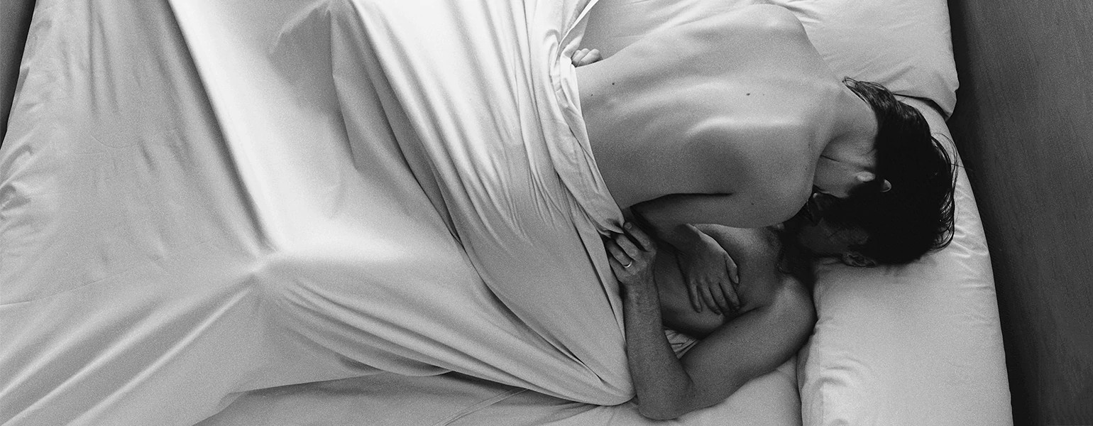A naked woman lays on top of a man, making love in bed under a white sheet.