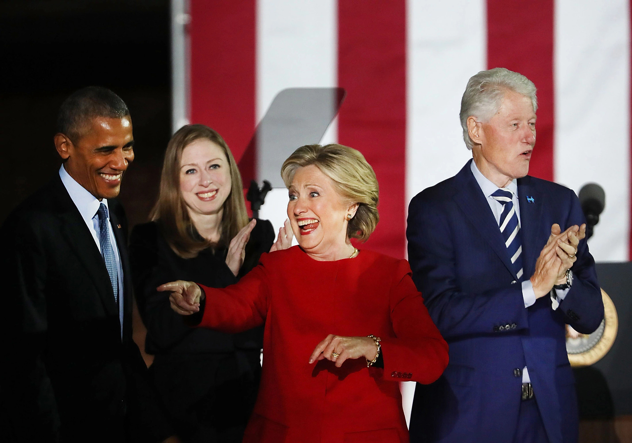 hillary clinton points off stage while barack obama, chelsea clinton and bill clinton stand behind her
