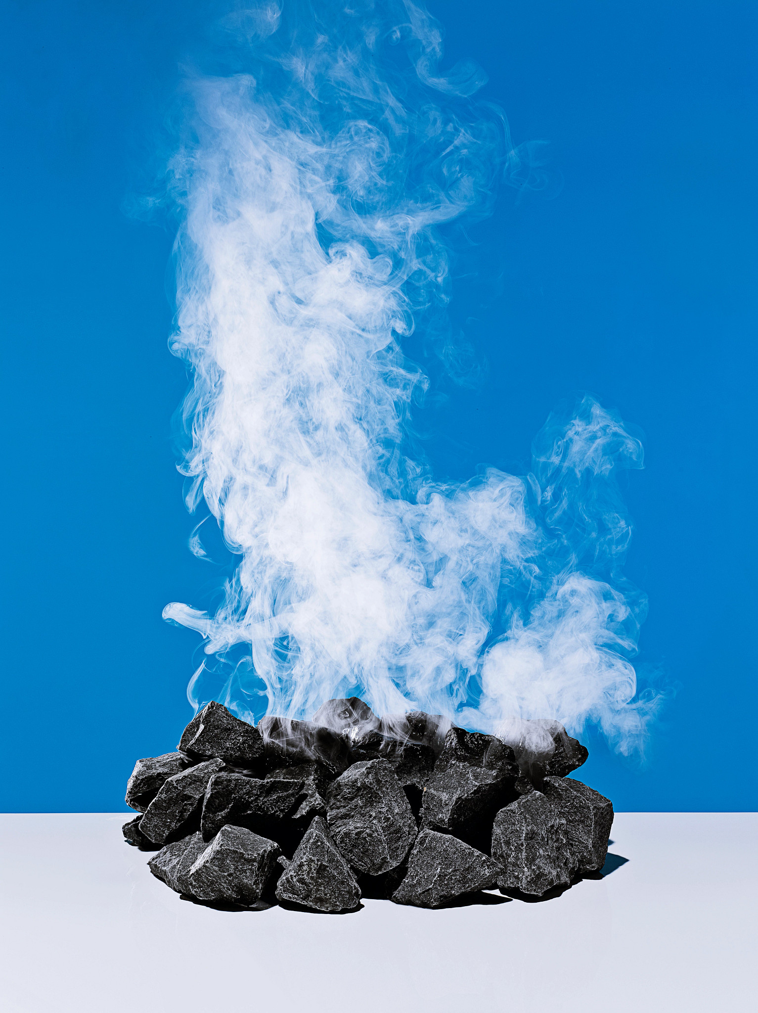 A pile of dark, textured sauna stones sits on a white surface with thick plumes of white steam rising against a solid bright blue background.