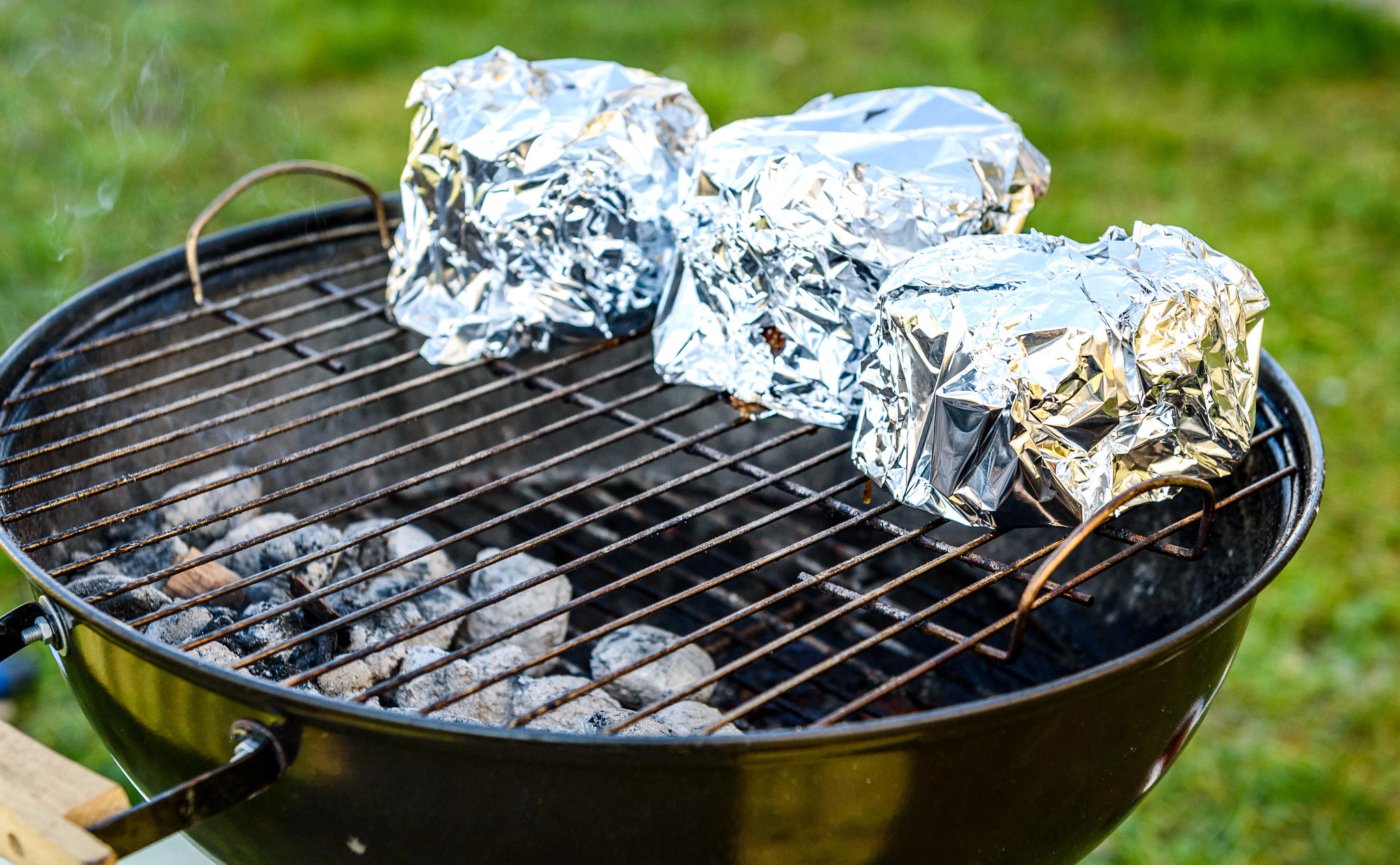Foil-wrapped food packets cooking on a charcoal barbecue grill outdoors with glowing coals beneath