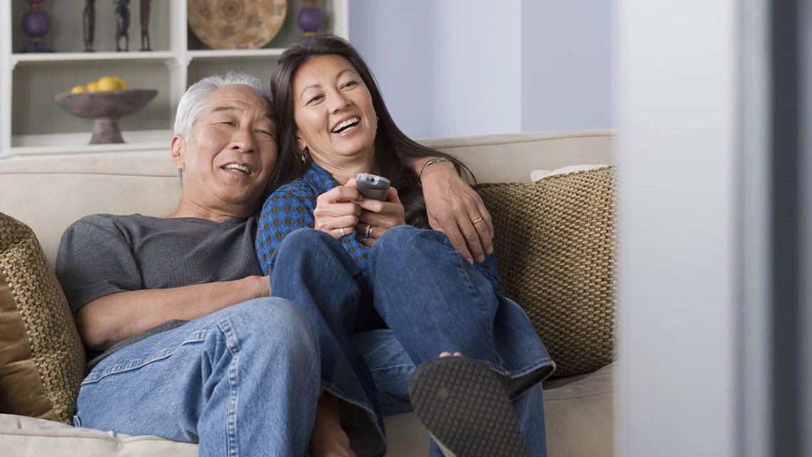 Asian couple curled up on couch together with remote laughing