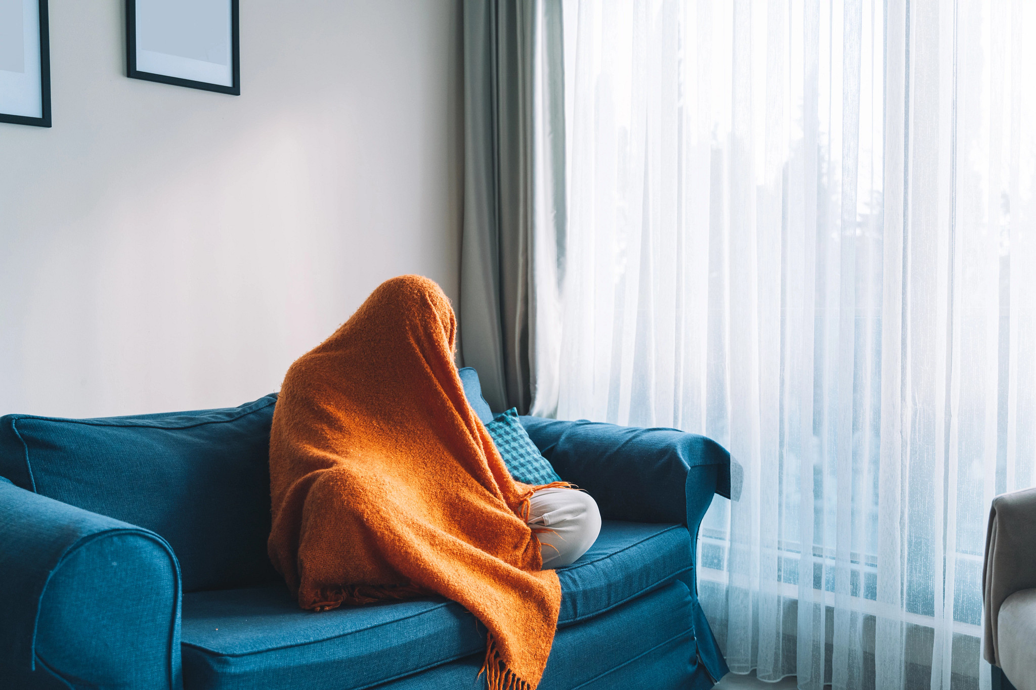 A person completely covered by an orange blanket sits on a blue couch next to a window.