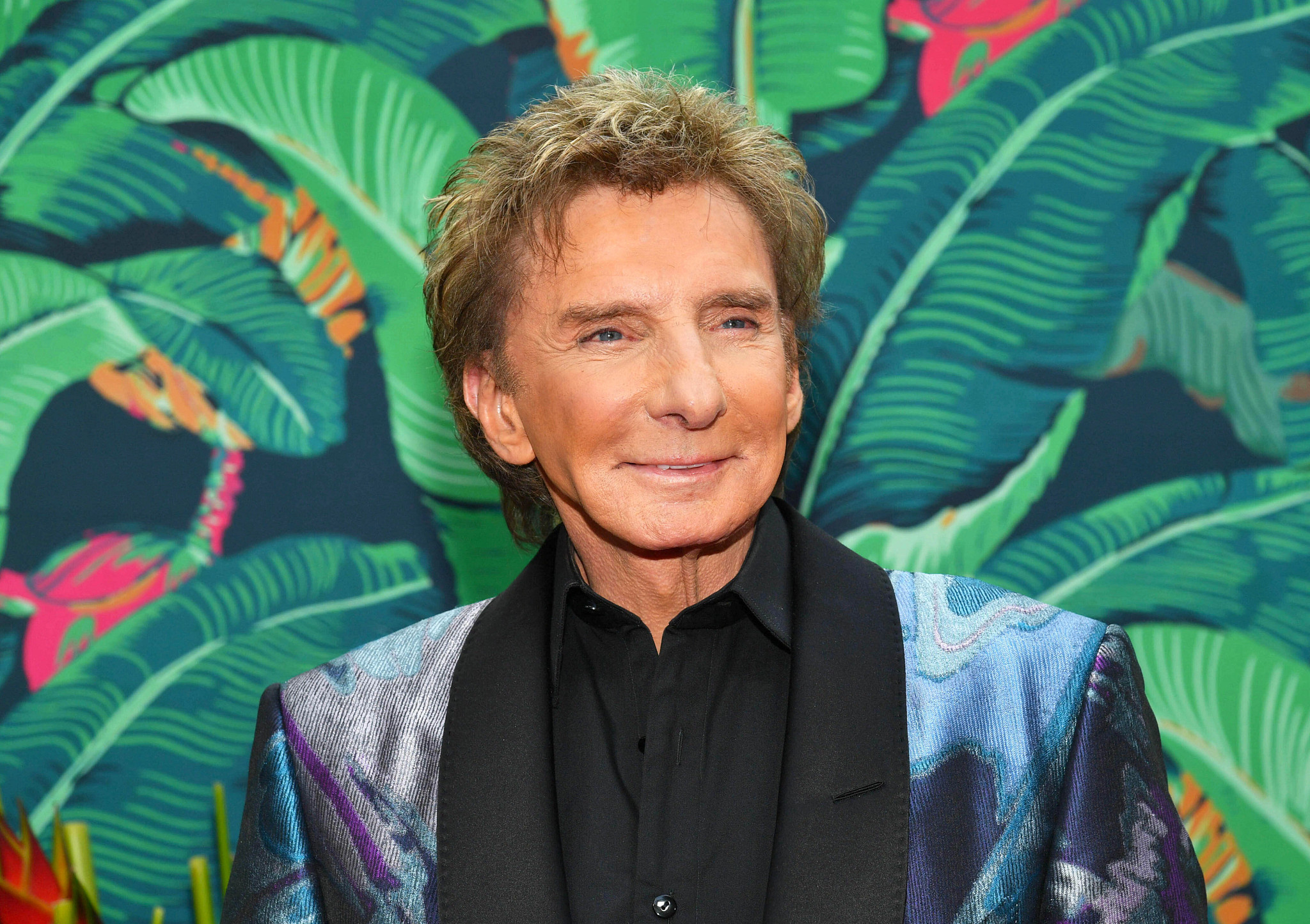 A headshot of singer Barry Manilow smiling, wearing a black collared shirt and a patterned blue and silver blazer against a background of green tropical leaves.