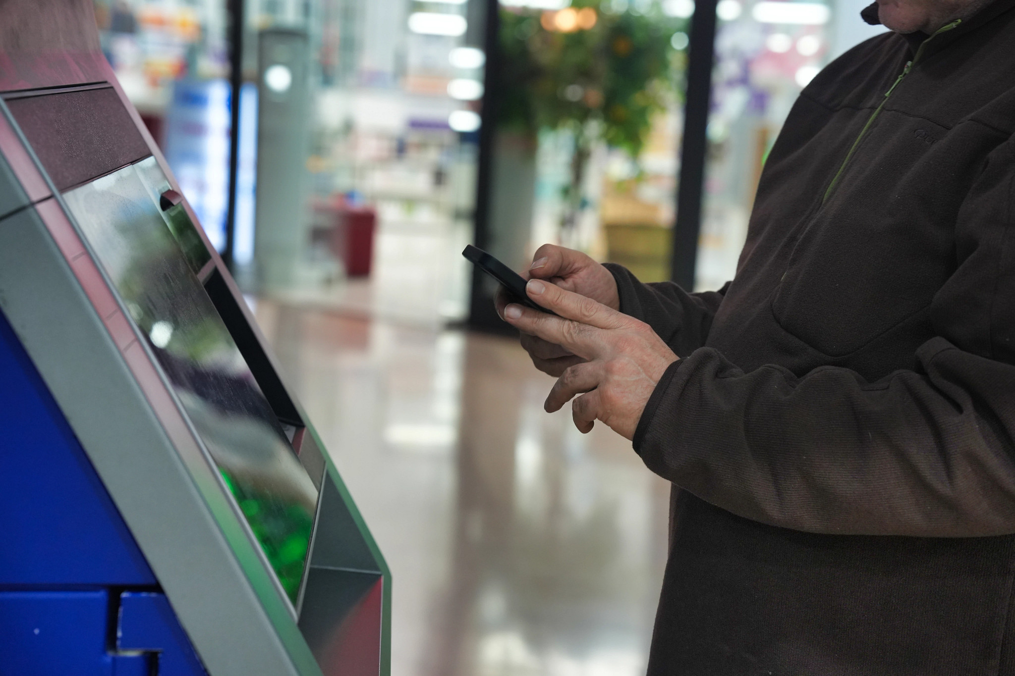 A man in a brown jacket holding a smartphone while interacting with a touchscreen kiosk in a public setting. 