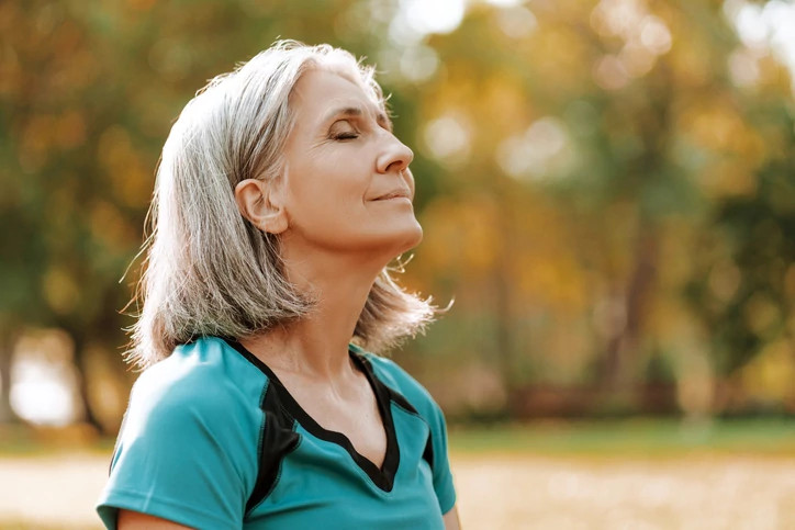 Senior woman wearing sportswear is enjoying a sunny autumn day in the park, closing her eyes and taking a deep breath of fresh air. Balance, harmony concept