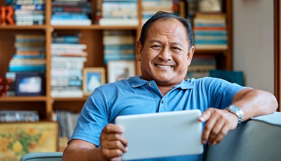 Man reading an e-Book on his tablet and smiling sitting on couch in front of large bookshelf filled with books.