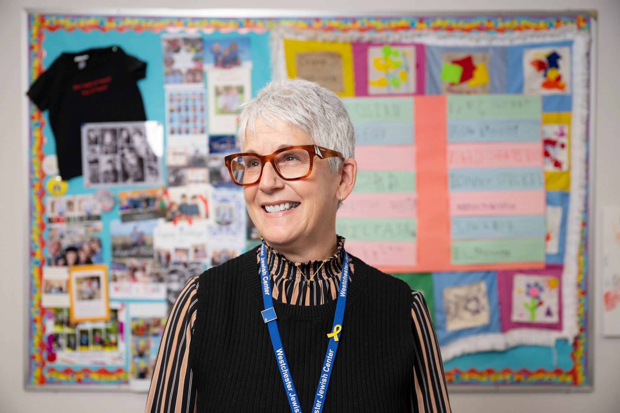 A closeup portrait of Ann Pircio smiling, wearing brown-framed glasses and a black sweater vest, standing in front of a bulletin board