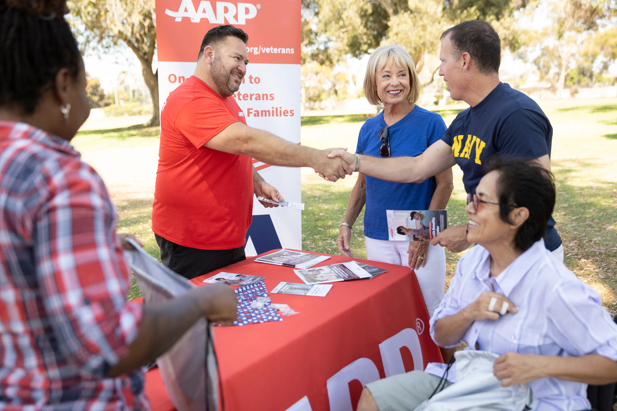 An AARP volunteer shakes hands with a military veteran and meets other visitors during a volunteer event for veterans and families at a park