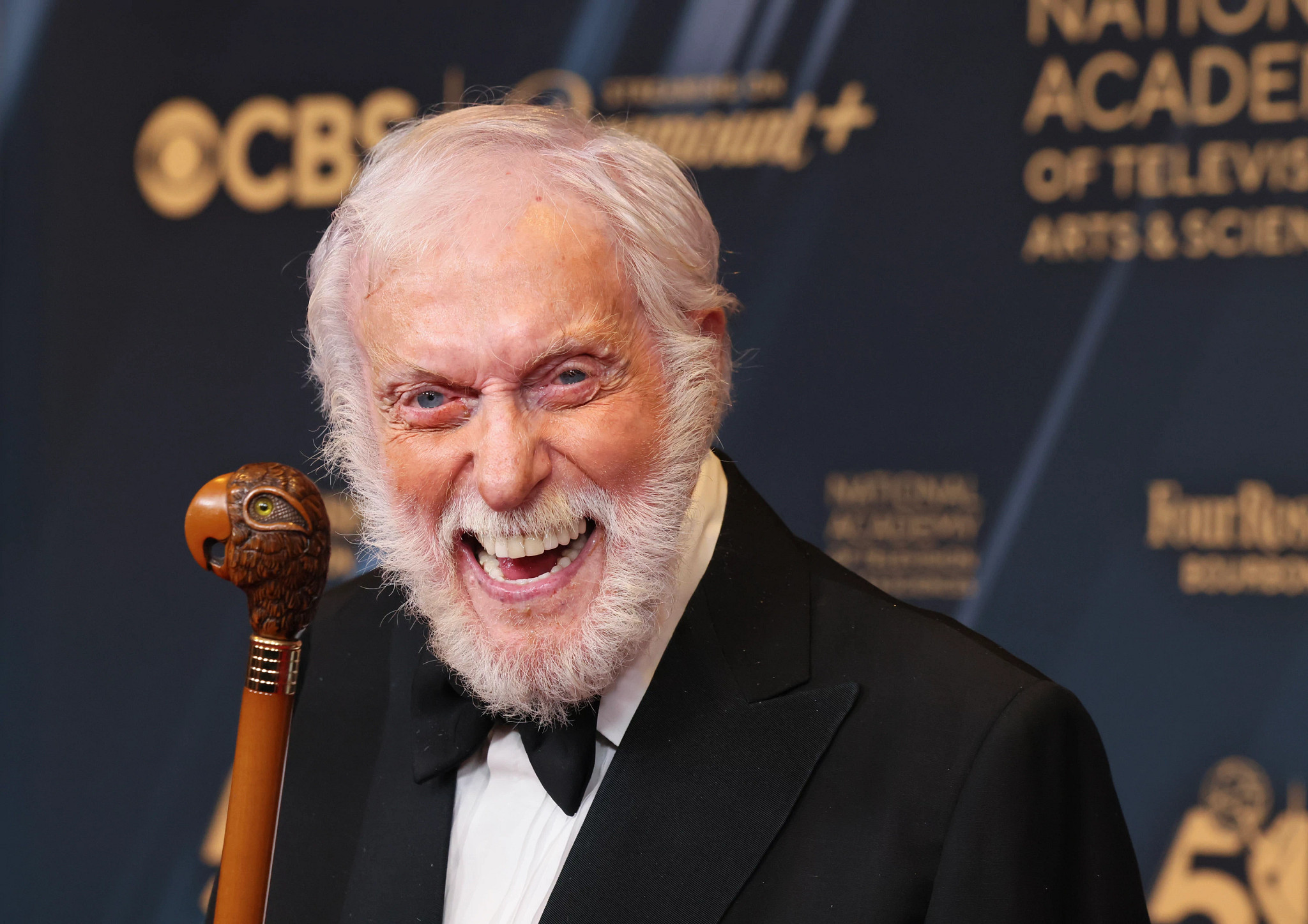 Dick Van Dyke, with an emphatically happy expression, wearing a suit and holding a wooden cane with a carved parrot head on the handle