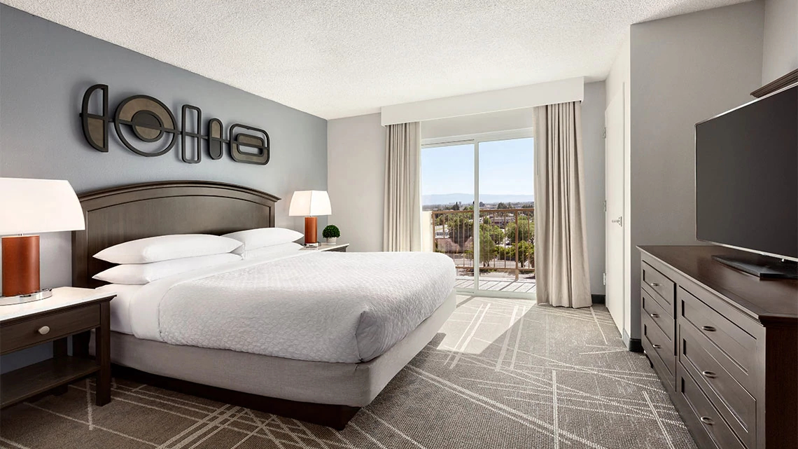 hotel room with king bed, white linens, gray and white carpet, balcony with sliding glass door, blue wall, two lamps, tv and dresser