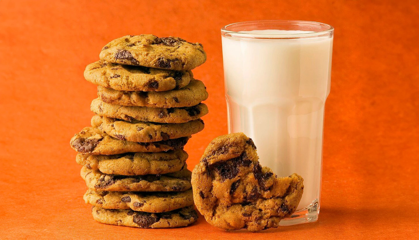 a stack of chocolate chip cookies is shown next to a glass of milk in front of an orange background