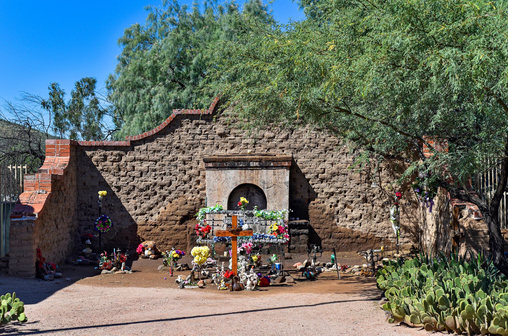El Tiradito a shrine with a cross, flowers and other personal items near a wall outside