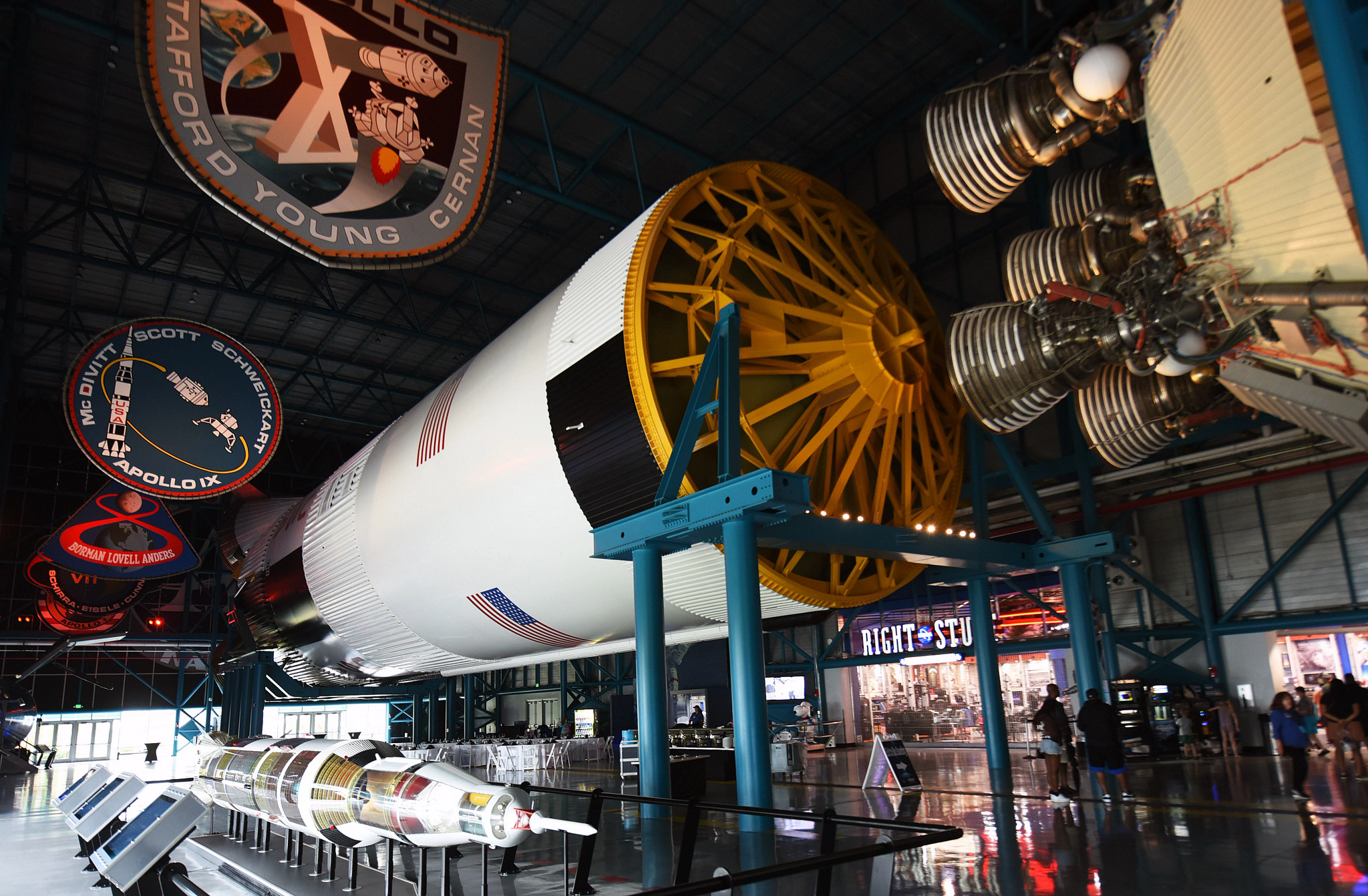 A large Saturn V rocket is displayed inside a space museum alongside Apollo mission exhibits and spacecraft hardware