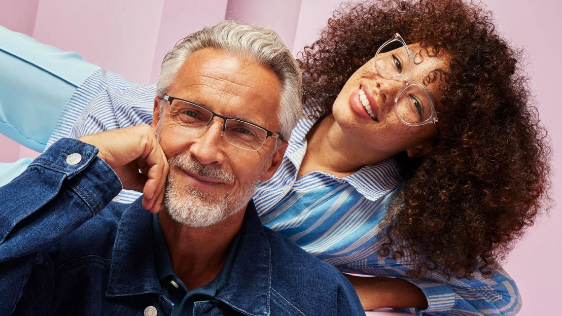 Target Optical Man sitting in front of a girl laying posed behind him. both smiling, both wearing denim blue clothes, both wearing glasses, pink background