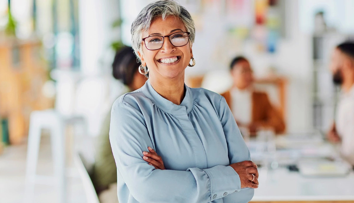 A smiling mature woman stands confidently in front of a blurred conference table with three people sitting at it.