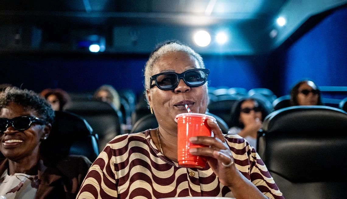 Movies for Grownups Mature woman drinking soda and wearing 3-D glasses in a dark movie theater with lights from the projector in the background