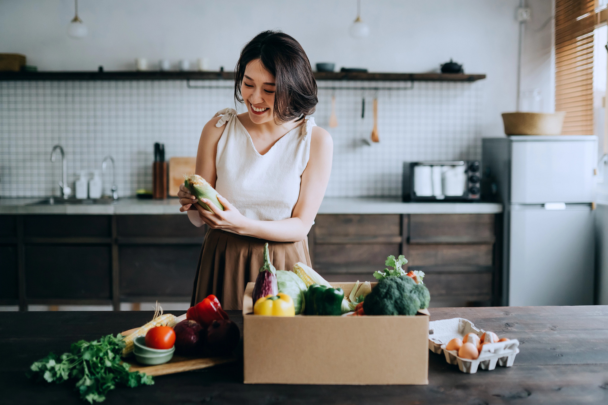 Beautiful smiling young Asian woman received a full box of colorful and fresh organic groceries ordered online by home doorstep delivery service.