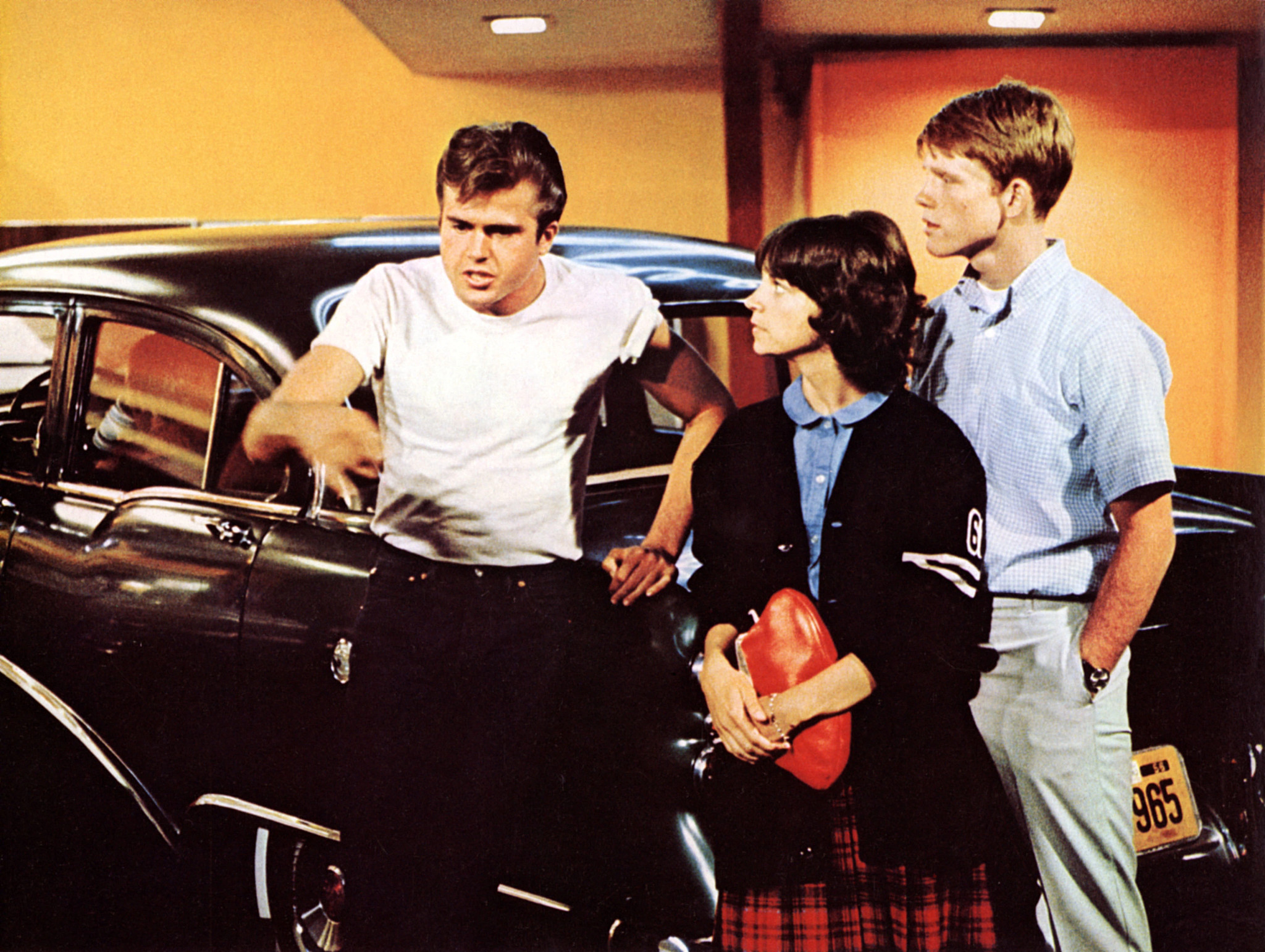Paul Le Mat, Cindy Williams and Ron Howard stand beside a vintage car in an indoor setting, engaged in conversation