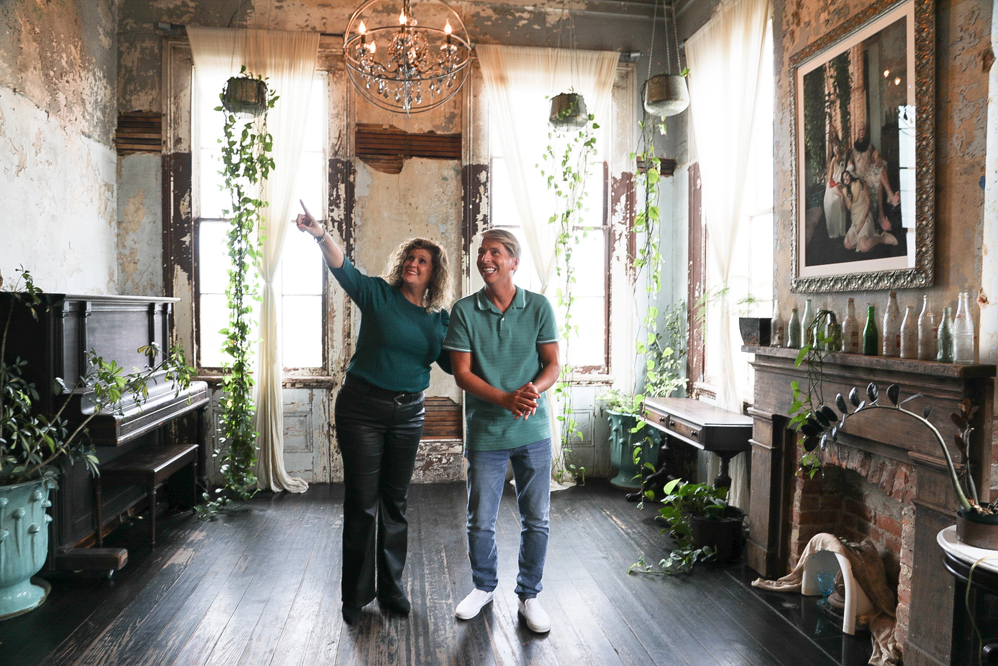 Aimee Ashe pointing to something, Jack McBrayer looking up at it inside a home with plants hanging from the ceiling and more on the floor, piano on the left, fireplace on the right