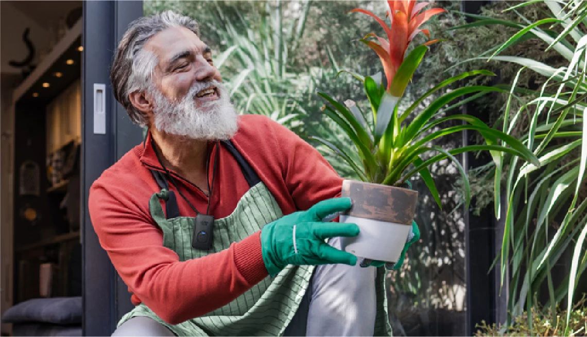 older man with gray and white hair, full beard, holding a potted plant/flower at a nursery or his home, wearing the around-the-neck alert device and gardening clothes including gloves, apron, long sleeves and pants