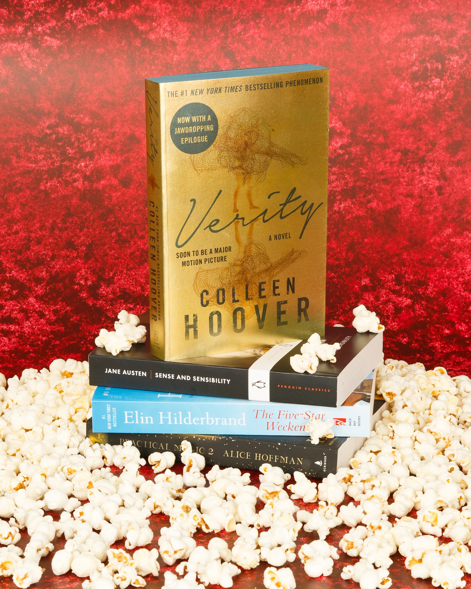 A styled book display featuring “Verity” by Colleen Hoover standing upright on a stack of novels with scattered popcorn against a vibrant red textured background