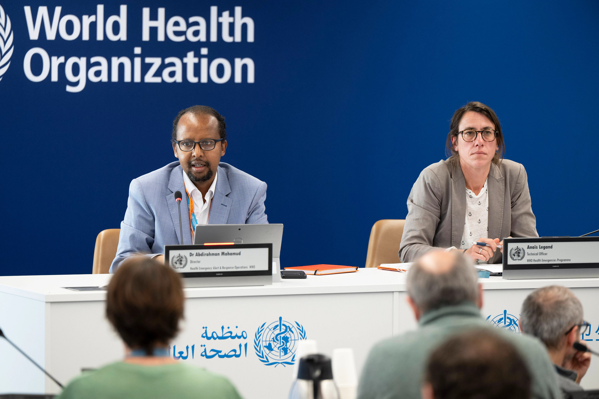 Two health officials speak at a panel during a World Health Organization news conference, seated at a white table with laptops and nameplates against a large blue WHO branded backdrop.