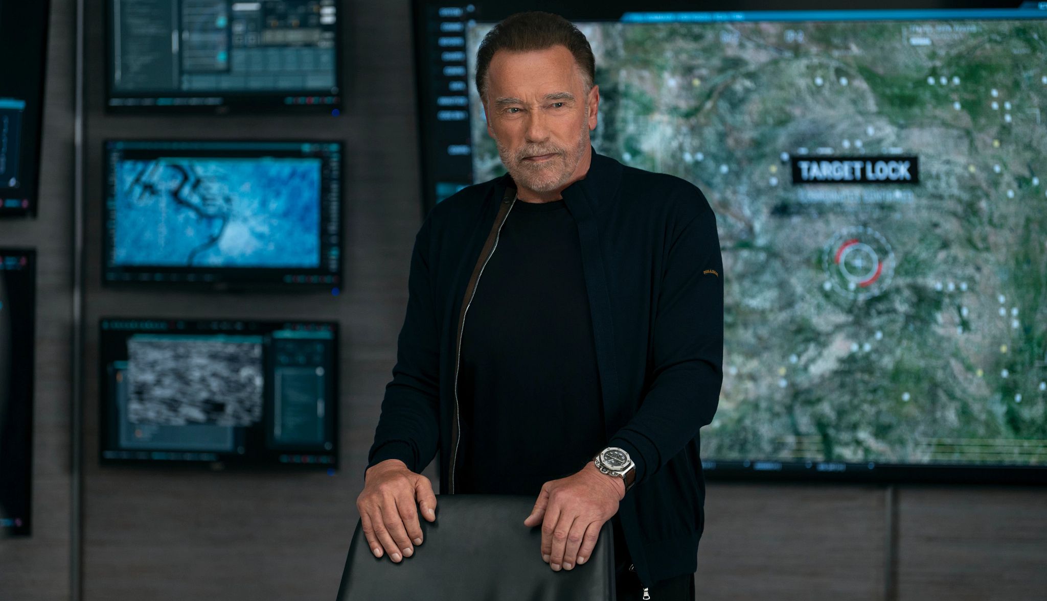 Netflix Summer TV Preview 2025 arnold schwarzenegger standing with his hands on the back of a black chair in a still image from the show fubar