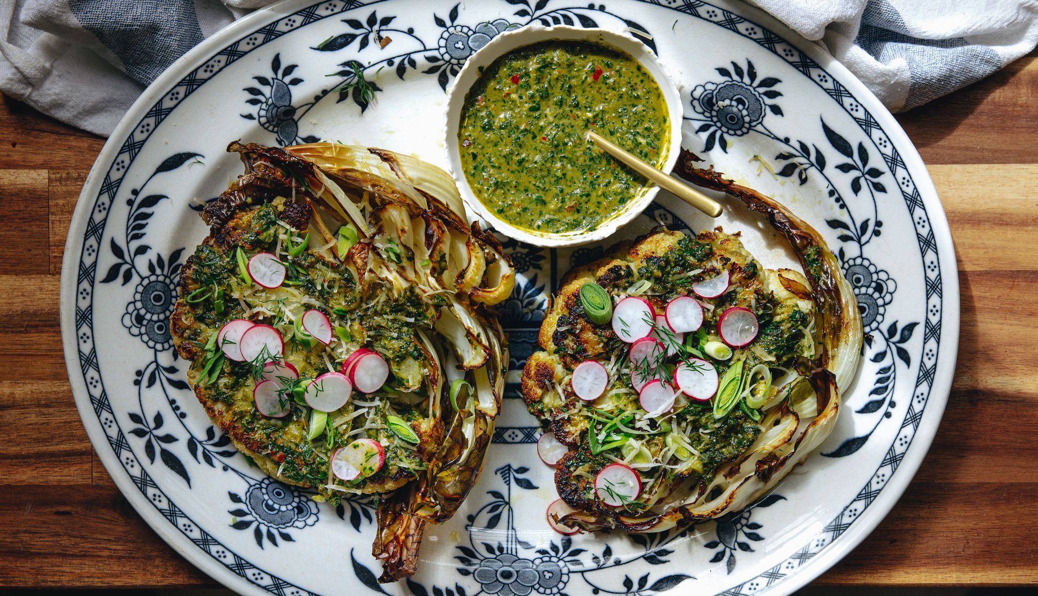 Cauliflower parmesan steaks with chimichurri Two cauliflower slices on a plate with chimichurri, radishes and scallions on them; small bowl of chimichurri next to them