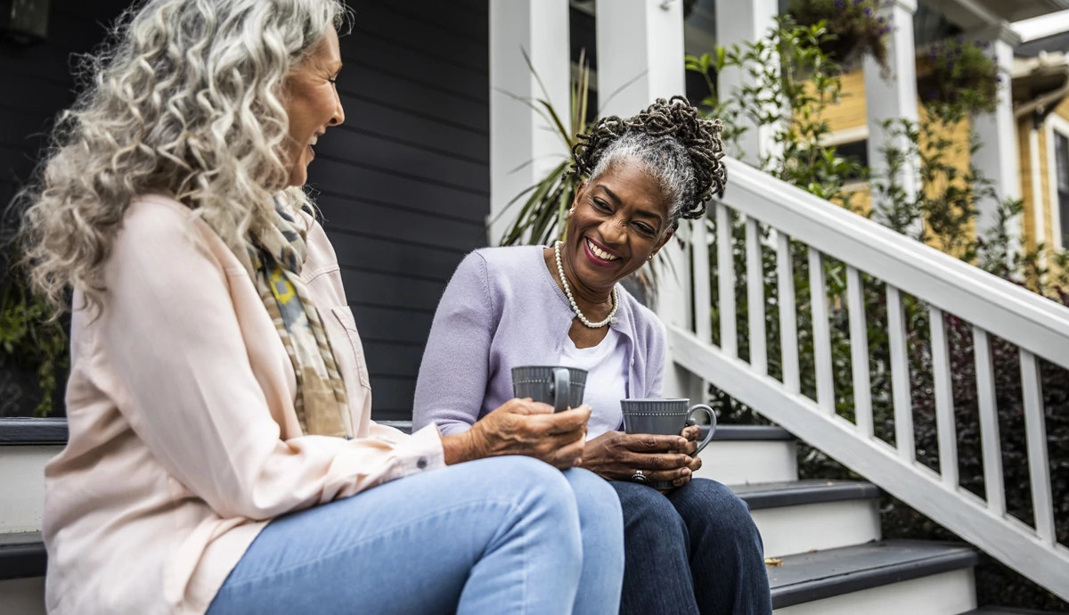 Two women sit on a front porch drinking coffee and laughing.
