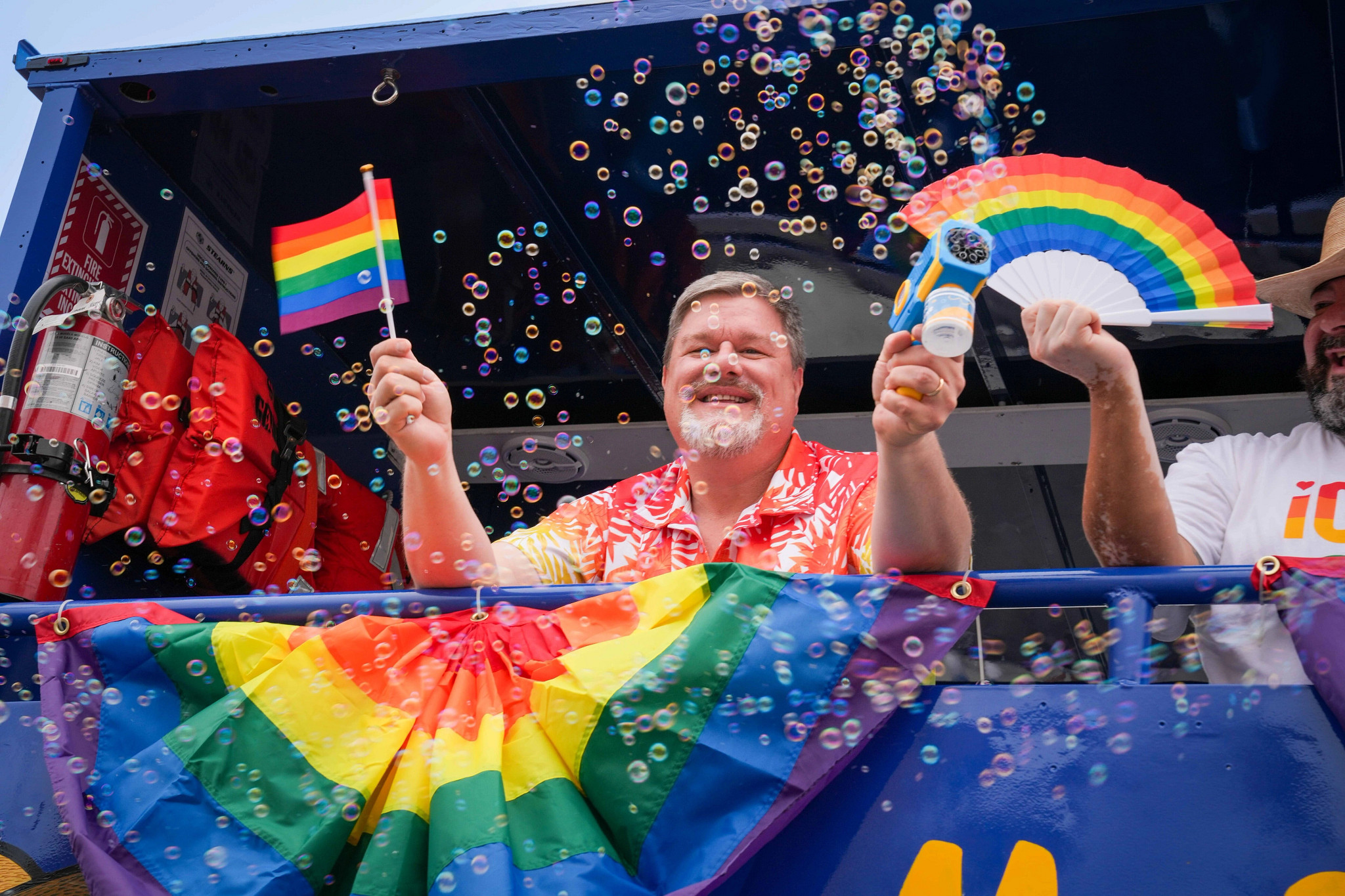A joyful man beams from a float decorated with rainbow flags, holding a mini Pride flag and a bubble blower.