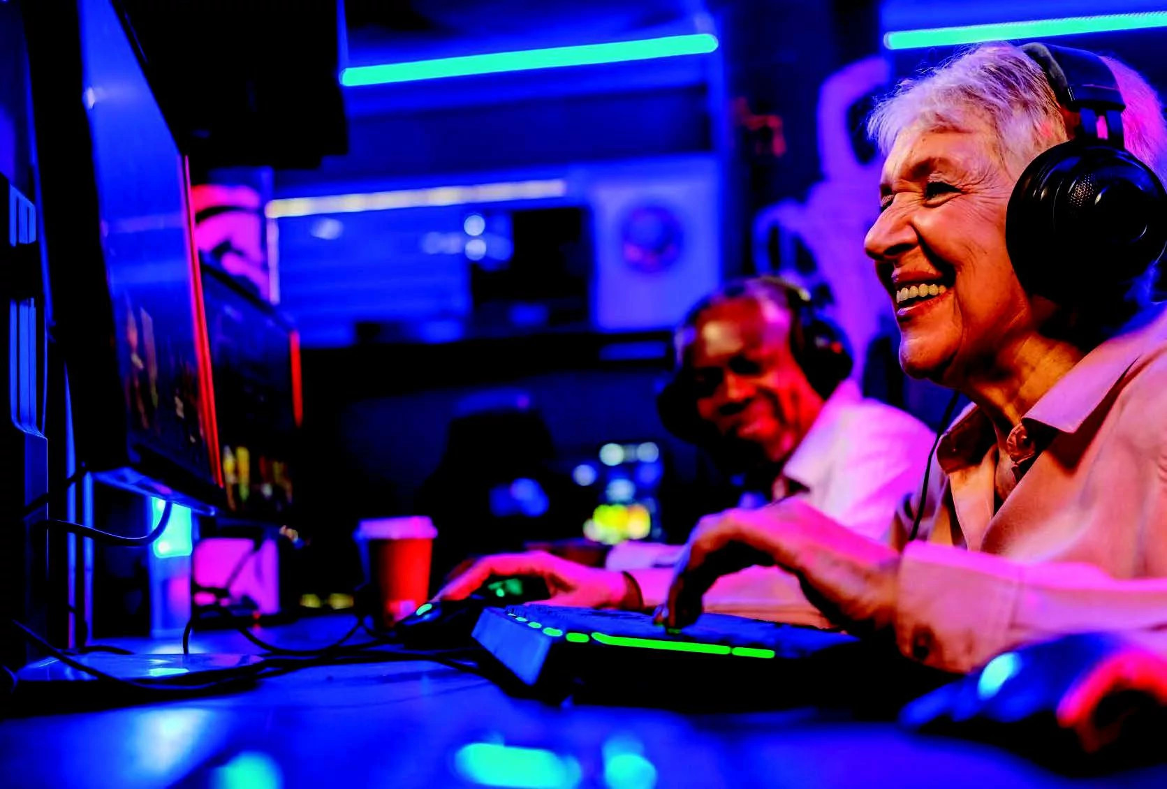 Older adults wearing headphones use computers in a colorful, neon-lit gaming room, with one woman smiling as she types on a keyboard