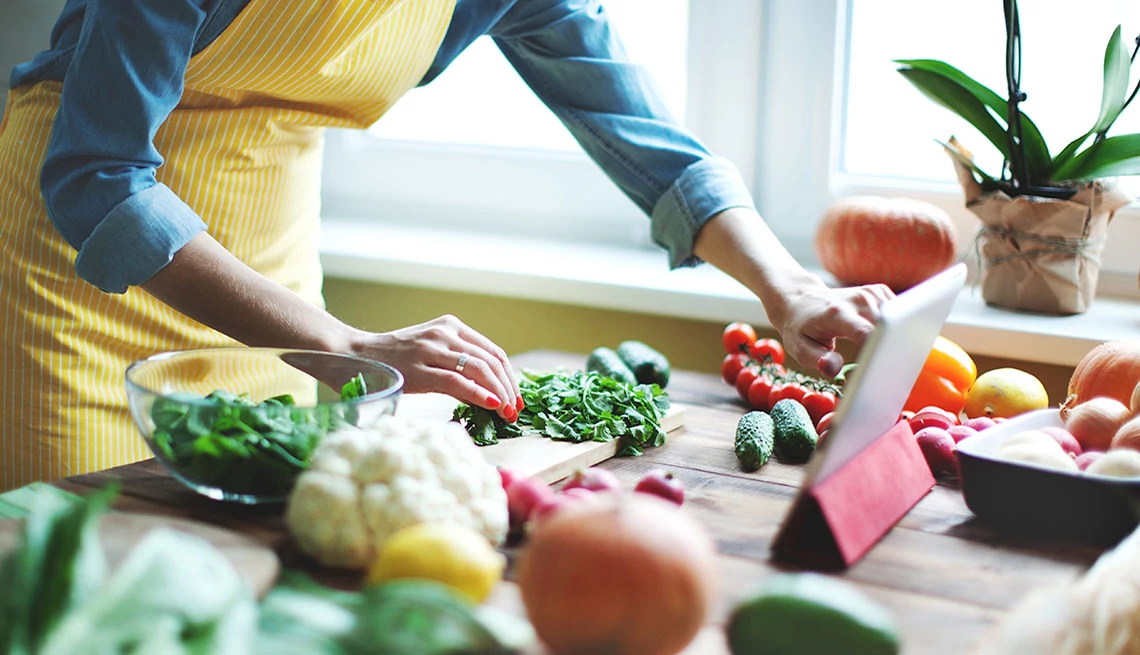AARP® Personalized Nutrition: Recipe Analyzer woman in yellow apron preparing vegetables looking at a tablet on counter