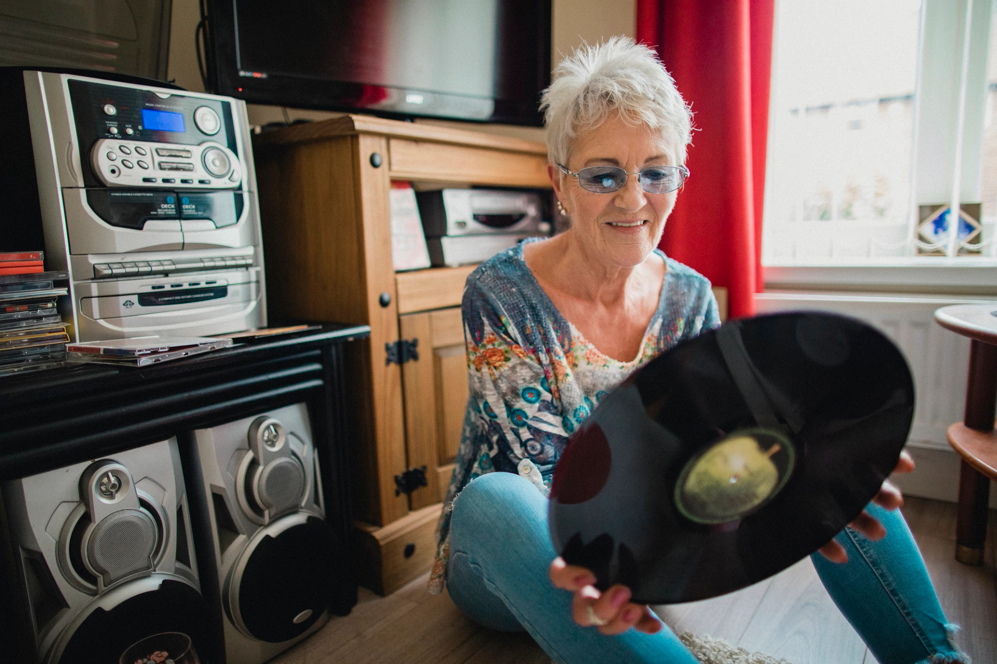 One woman sitting on the floor in her living room, holding a vinyl record in her hands and listening to music from a record player.